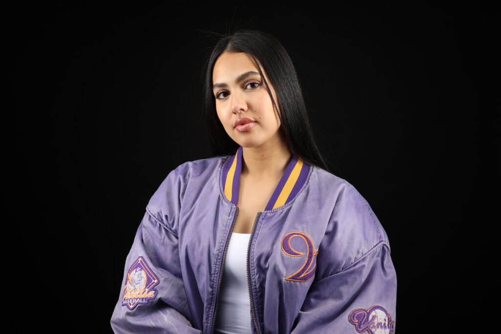 A promotional photo of Manal. She is wearing a purple jacket against a black background, looking straight at the camera.
