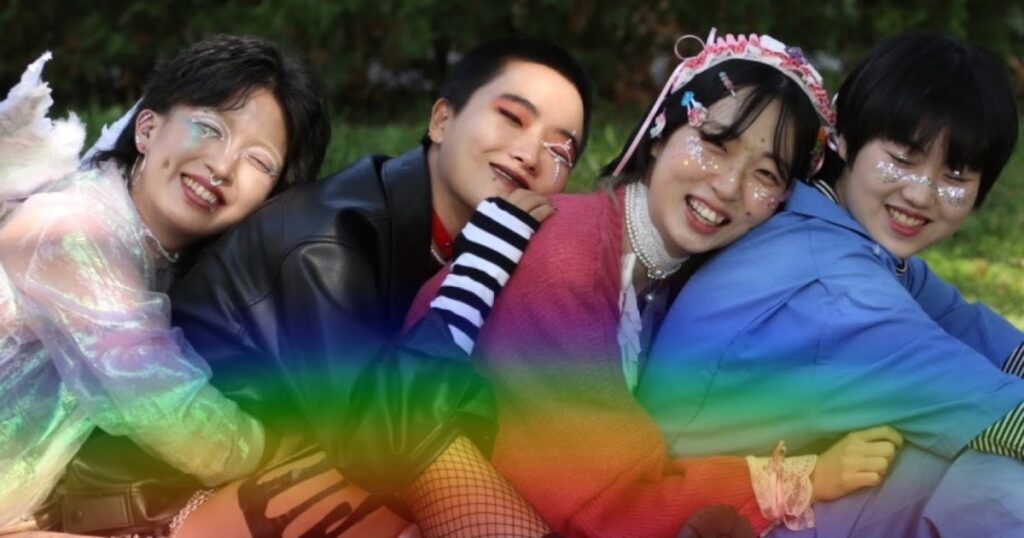 A photo of the members of QI.X in a line, hugging each other while looking into the camera. A rainbow is superimposed along the bottom of the photo.