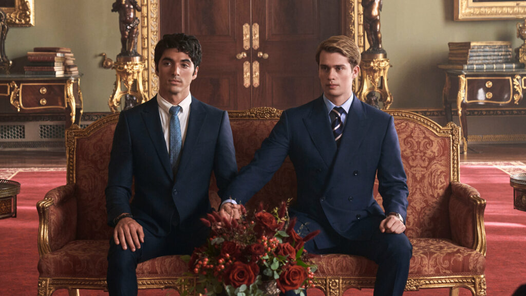 A photo still from Red, White, and Royal Blue. Alex and Henry are seated next to each other on a couch, wearing suits, holding hands and looking off camera.