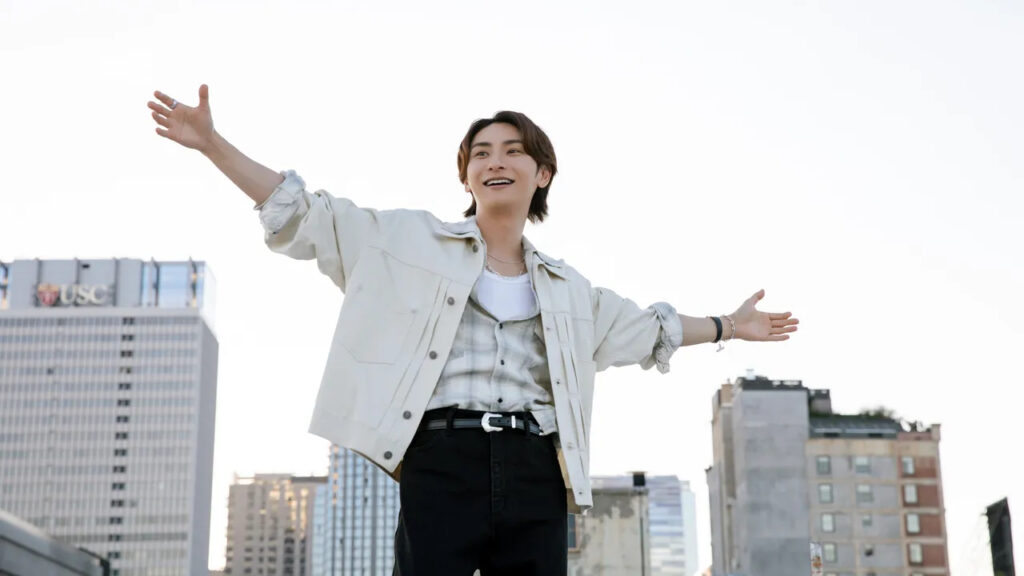 A promotional image of Shinjiro. He is standing outside with the camera near the ground looking up at him. His arms are outstretched to his sides, and he is looking off to the left with a smile on his face.