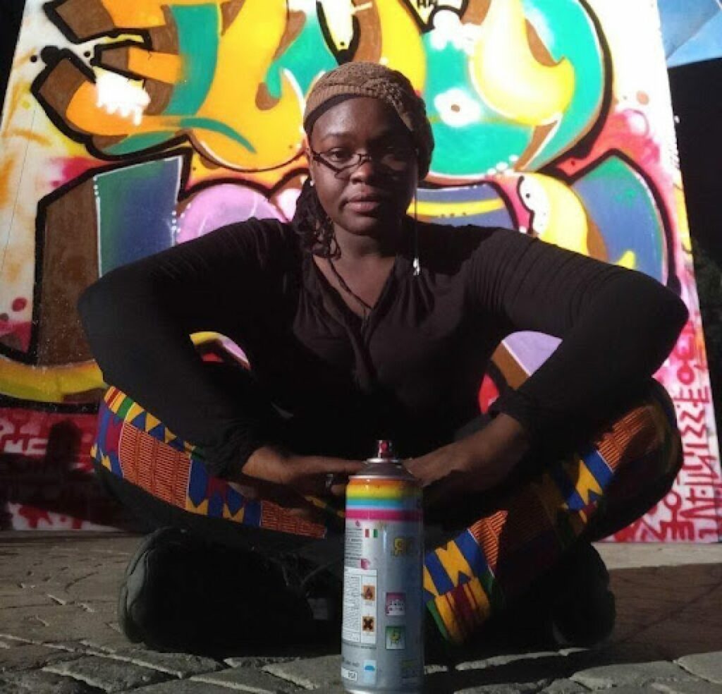 A photo of Zeinixx sitting cross legged in front of one of her pieces. A spray can sits directly in front of her.