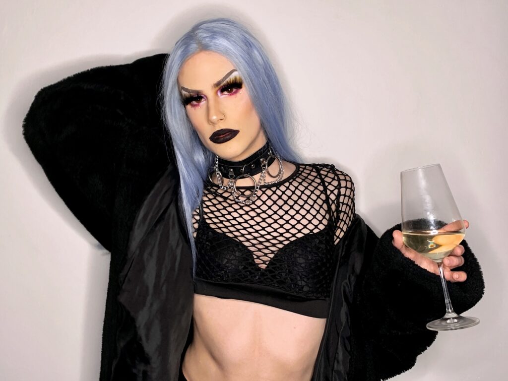 A photo of DIMA wearing a black crop top with black mesh around the neck and a blue wig. She is holding a glass of wine and posing directly into the camera.