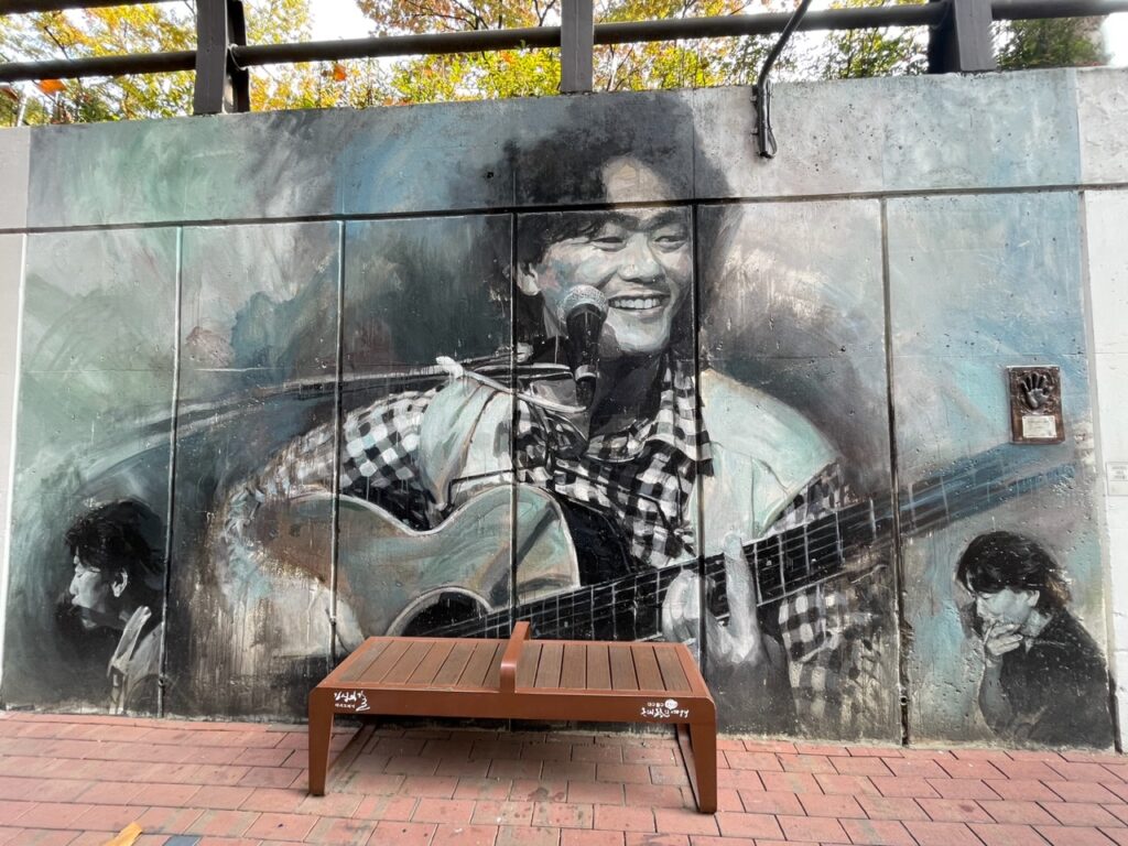 A photo of a mural on Kim Kwang-seok Street. The mural shows an image of Kim Kwang-seok smiling and holding a guitar, with a microphone in front of his face. There is a brown bench against the wall in front of the mural.