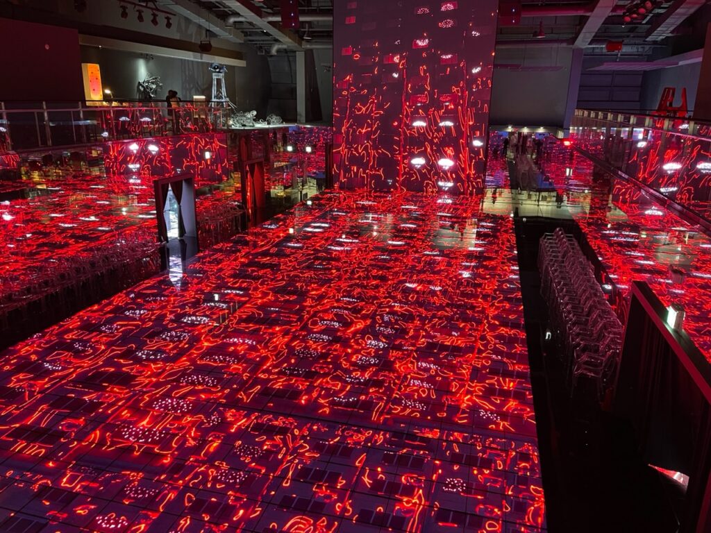 A photo of a digital artwork by Kim Yong Min. It shows a large room where the floor and walls are all screens, and an image of red patterns is projected on all the screens.
