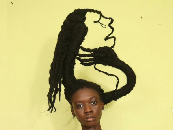 A photo of a work of hair art by Laetitia Ky. A woman's head is seen at the bottom of the frame, and above her, her hair creates a profile of a woman against a yellow background.