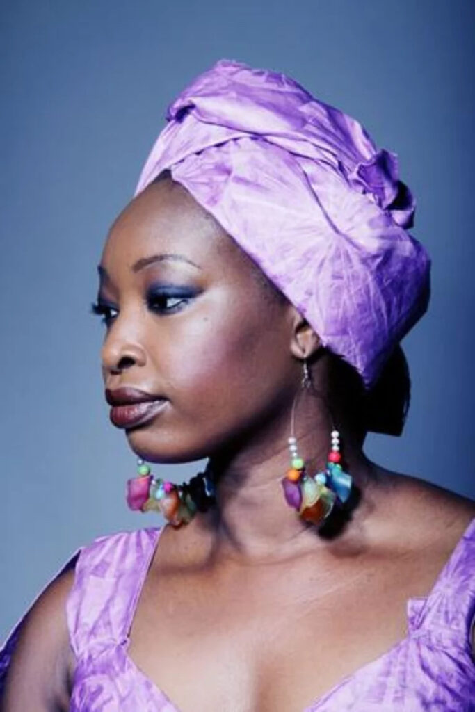 A profile portrait of Mounira Mitchala. She is wearing a purple dress with a purple headscarf and large dangling earrings.
