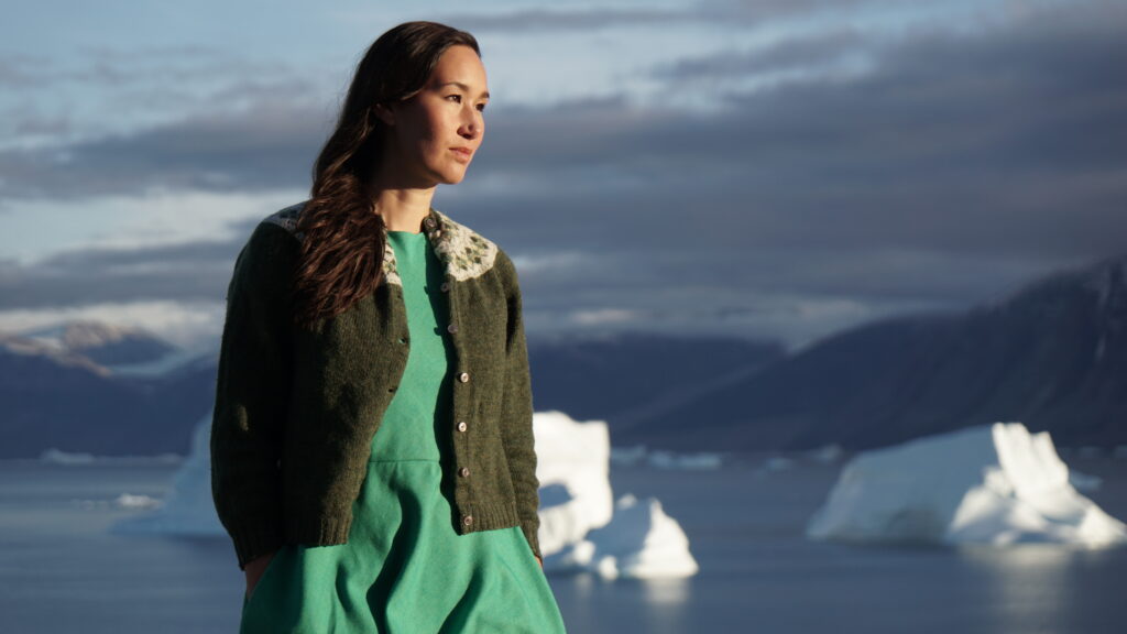 A photo of Nive Nielsen. She is standing outside, bathed in sunlight, with the ocean with icebergs in the background. She is looking off to the right of the camera.