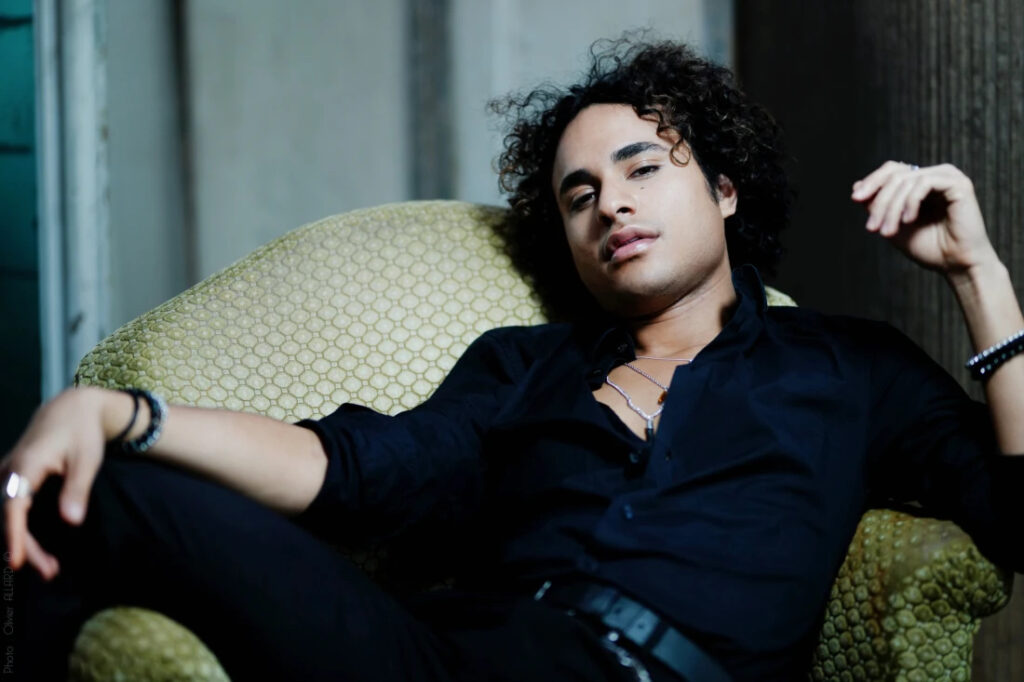 A photo of Samuel Mariño posed reclining on an armchair, looking sultry into the camera.